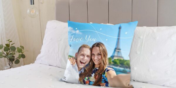 A custom photo cushion on a bed. The cushion features a picture of a smiling couple, with a man on the left embracing a woman on the right. Both are looking directly at the camera. Behind them, the Eiffel Tower is visible against a blue sky with some white decorative swirls and small hearts, and the words "Love You" written in a stylised font.