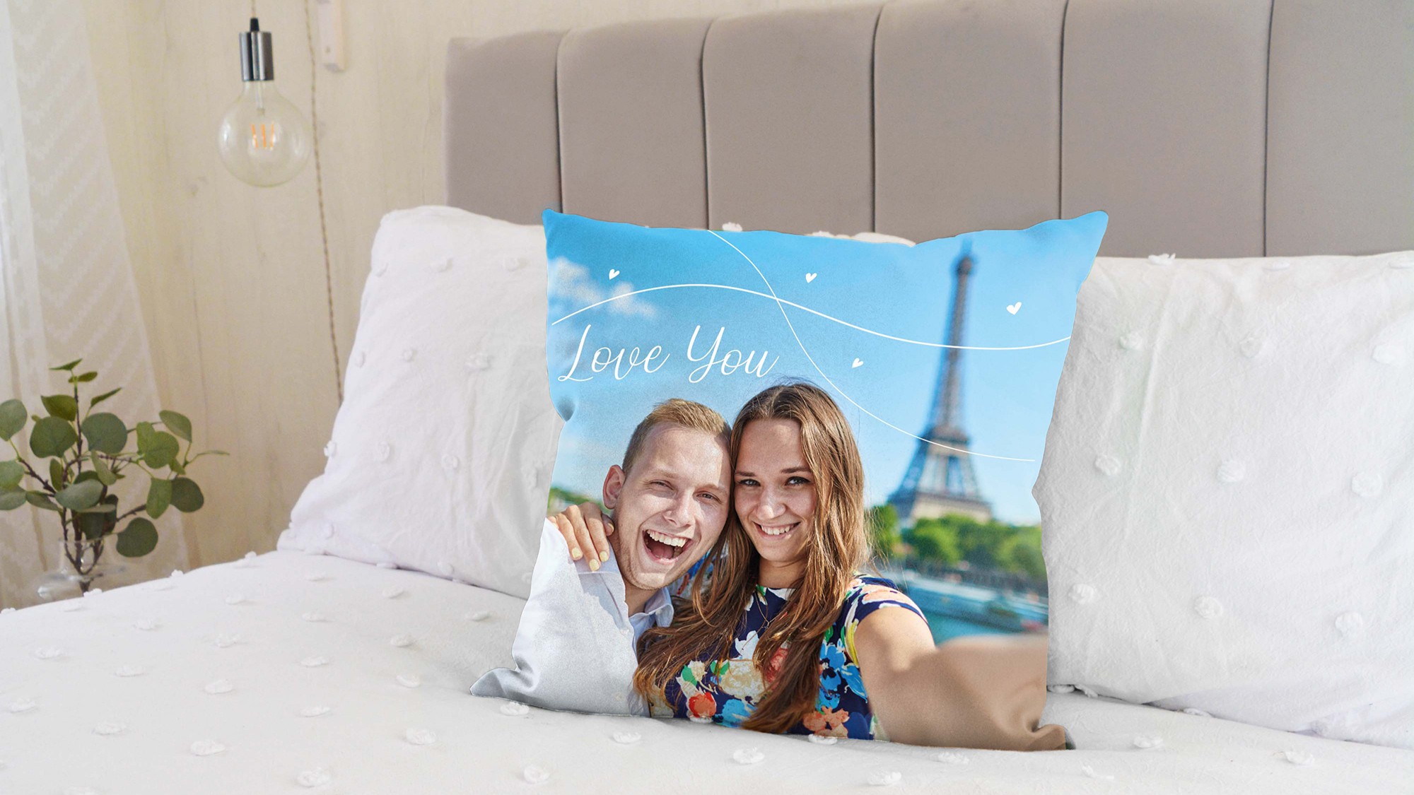 A custom photo cushion on a bed. The cushion features a picture of a smiling couple, with a man on the left embracing a woman on the right. Both are looking directly at the camera. Behind them, the Eiffel Tower is visible against a blue sky with some white decorative swirls and small hearts, and the words "Love You" written in a stylised font.