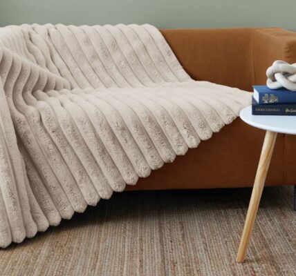 A cosy living room scene featuring a light brown, two-seater sofa. A plush, cream-coloured ribbed faux fur blanket is draped over the sofa, cascading down the front