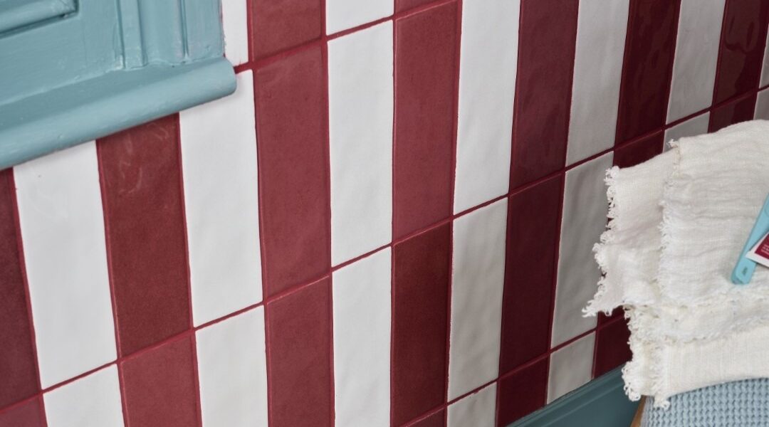 A wall is tiled with alternating vertical stripes of glossy, brick-red and off-white rectangular tiles. The tiles are arranged in a staggered pattern, with each row offset from the one below it. The grout between the tiles is a reddish-brown colour.