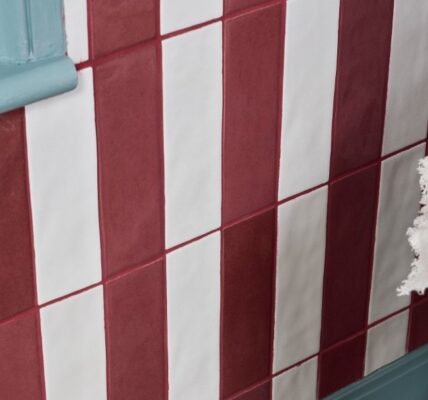 A wall is tiled with alternating vertical stripes of glossy, brick-red and off-white rectangular tiles. The tiles are arranged in a staggered pattern, with each row offset from the one below it. The grout between the tiles is a reddish-brown colour.