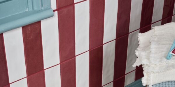 A wall is tiled with alternating vertical stripes of glossy, brick-red and off-white rectangular tiles. The tiles are arranged in a staggered pattern, with each row offset from the one below it. The grout between the tiles is a reddish-brown colour.