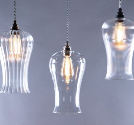 A trio of glass pendant lights hangs against a soft grey background. The lights are suspended by braided fabric cords. The two outer fixtures are identical, featuring a ribbed, fluted glass shade that flares out at the bottom. The central light has a smooth, clear glass shade with a similar bell-like shape.