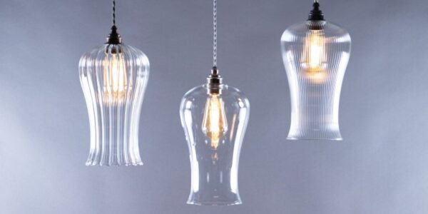 A trio of glass pendant lights hangs against a soft grey background. The lights are suspended by braided fabric cords. The two outer fixtures are identical, featuring a ribbed, fluted glass shade that flares out at the bottom. The central light has a smooth, clear glass shade with a similar bell-like shape.