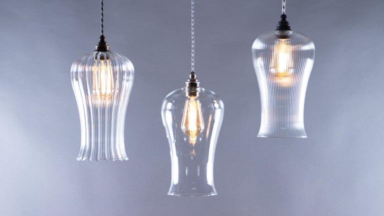 A trio of glass pendant lights hangs against a soft grey background. The lights are suspended by braided fabric cords. The two outer fixtures are identical, featuring a ribbed, fluted glass shade that flares out at the bottom. The central light has a smooth, clear glass shade with a similar bell-like shape.