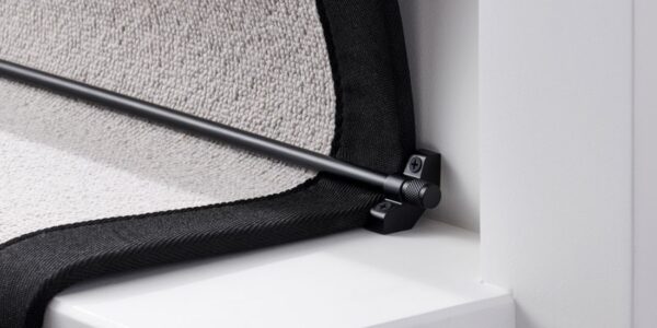 A close-up, low-angle shot of a black stair carpet runner rod installed on a white staircase. The black runner rod is held in place by a matching black bracket on the left, which is screwed into the white stair riser. The bracket has a decorative knob on the front, and the screw is visible.
