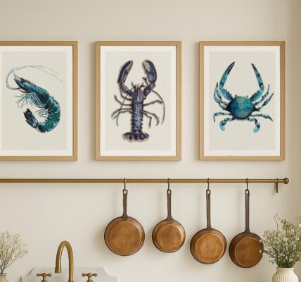 A set of three framed art prints depicting shellfish, including a shrimp, a lobster, and a crab, are displayed in a row on a light-coloured wall above a kitchen counter. Below the prints, there's a brass rail with four copper pans hanging from hooks.