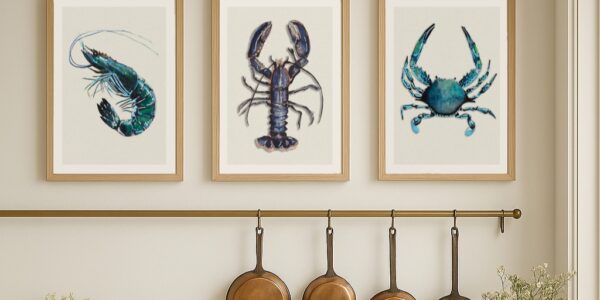 A set of three framed art prints depicting shellfish, including a shrimp, a lobster, and a crab, are displayed in a row on a light-coloured wall above a kitchen counter. Below the prints, there's a brass rail with four copper pans hanging from hooks.
