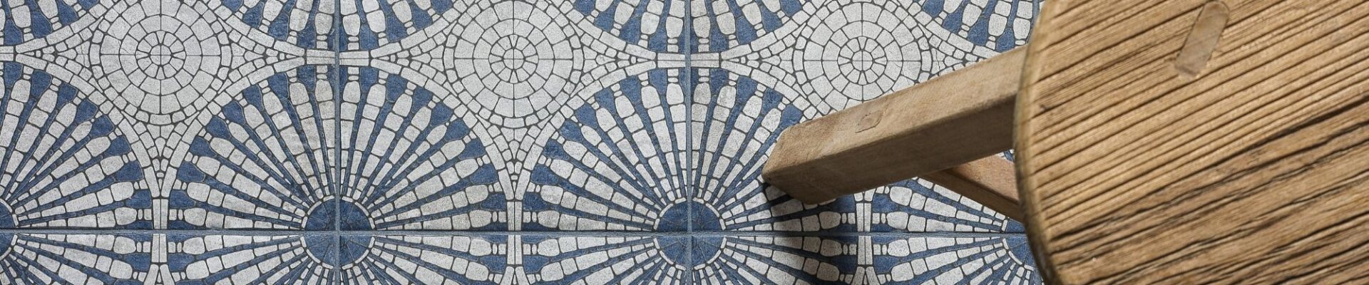 A bird’s eye view of a patterned floor tile. The light grey and blue tile has a circular geometric pattern. A small wooden stool is also seen.