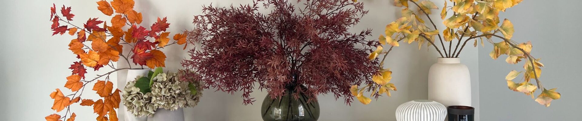 A white marble fireplace is decorated with three vases of artificial autumnal leaves. A small bunch of realistic-looking grey-green hydrangeas sits next to the vase on the far left. The vase on the left holds bright orange maple leaves on a branch. The centre vase holds a sprawling, deep burgundy-coloured branch with fine, fern-like leaves.