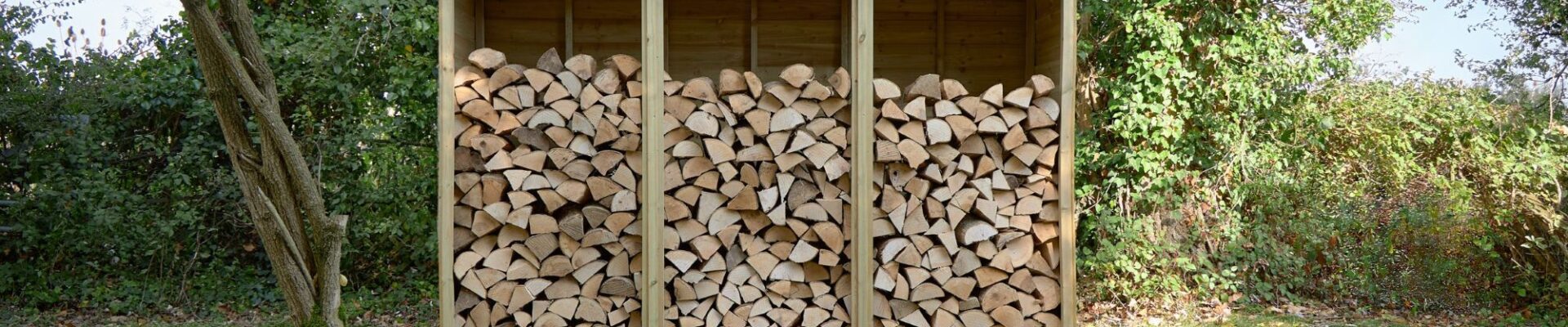A three-bay wooden log store is filled with neatly stacked firewood. It sits on a patch of grass in front of a leafy green hedge, with trees visible on either side and in the background. The store has a sloped roof that overhangs the front.
