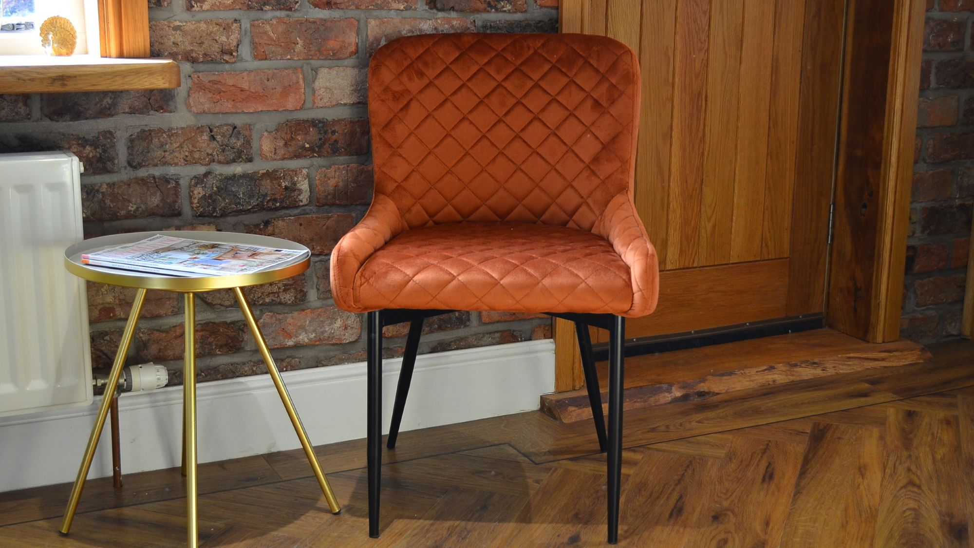 A quilted burnt orange velvet chair with black metal legs, sits near a small, round side table with a silver top and gold-coloured, tripod-style legs. A rustic brick wall behind the chair.