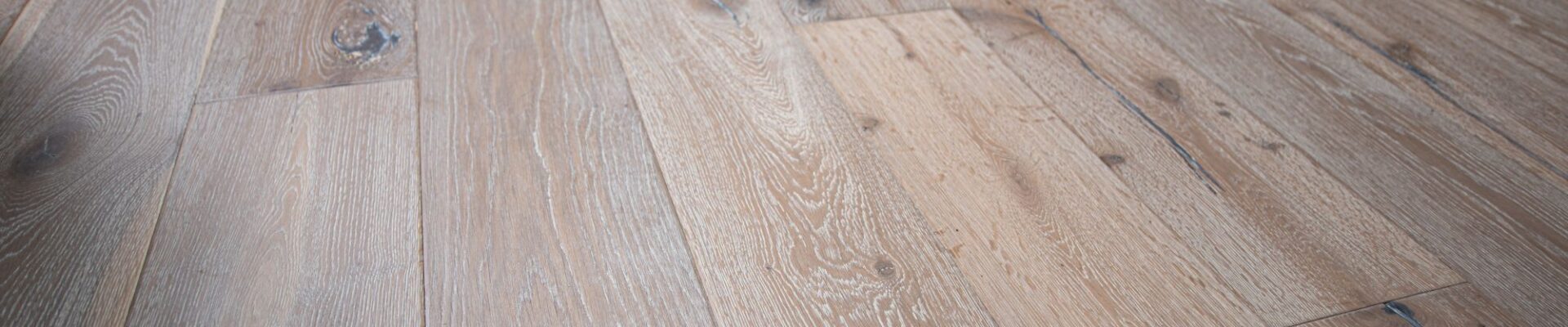 Distressed, light grey engineered oak flooring with visible wood grain and dark knots. The wide planks are laid parallel to each other, running toward a pale blue wall and a wooden cabinet. The lighting highlights the texture and varied tones of the floor.