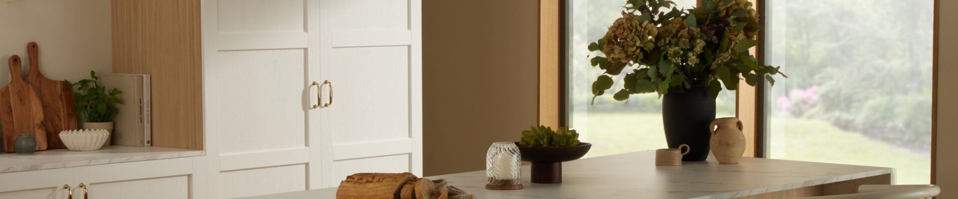 A bright kitchen island featuring a white marble countertop with grey veining and a light wood base. Two light wood and woven rush wishbone chairs are tucked underneath. Above the island, three fluted clear glass pendant lights hang.