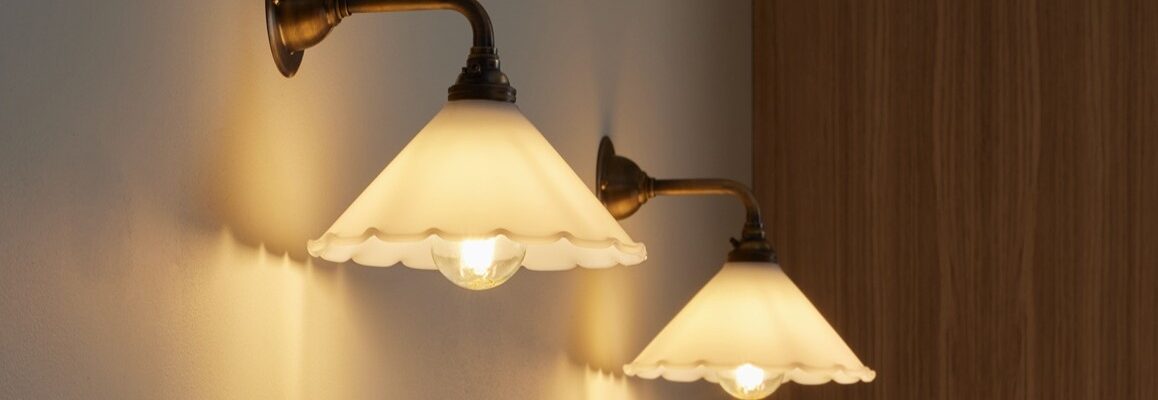 A wall light which features a frilled, scalloped edge, a cone Shape and an antique finish. The arm and wall plate have a warm, aged metal look.