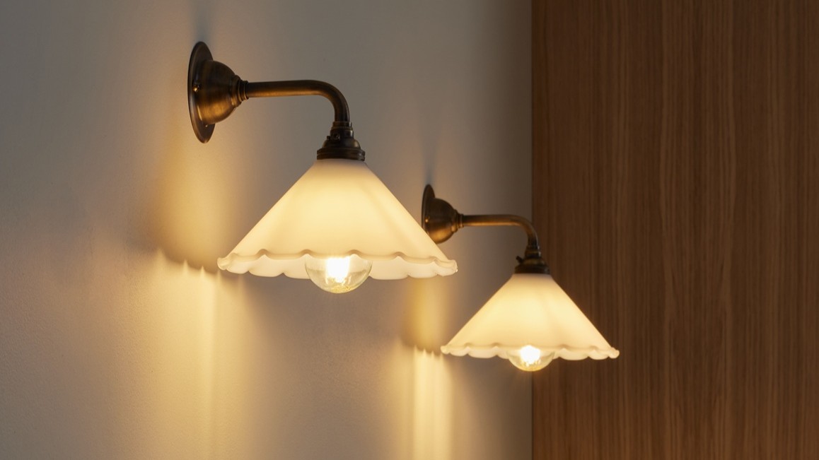 A wall light which features a frilled, scalloped edge, a cone Shape and an antique finish. The arm and wall plate have a warm, aged metal look.