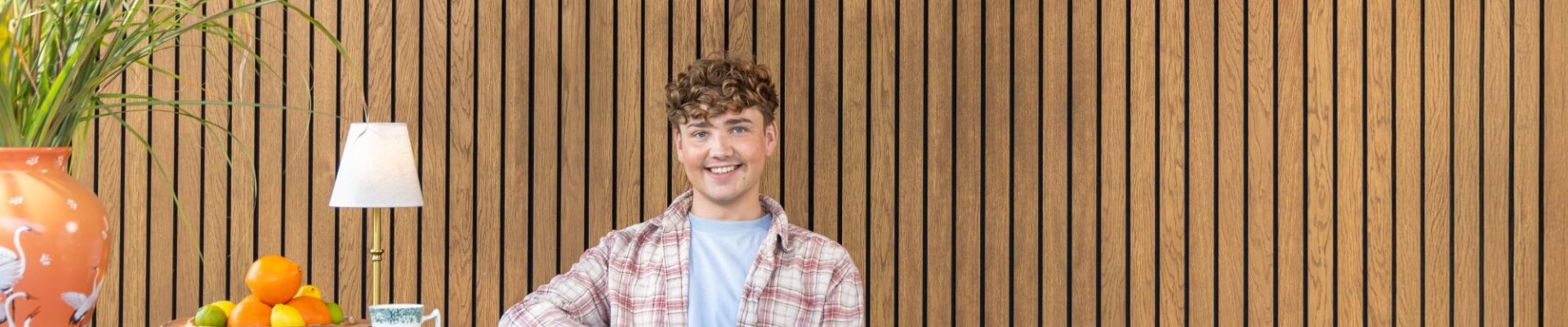 Interior Designer Jack Kinsey sits in front of a slatted wall. The slatted wall is very rich in colour and brings warmth to the image.