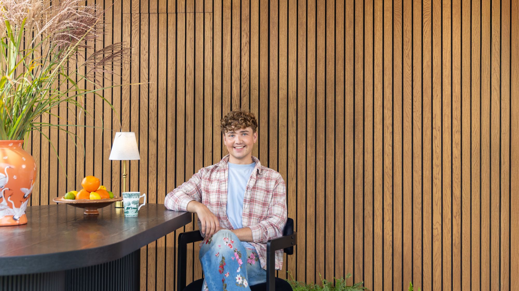 Interior Designer Jack Kinsey sits in front of a slatted wall. The slatted wall is very rich in colour and brings warmth to the image.