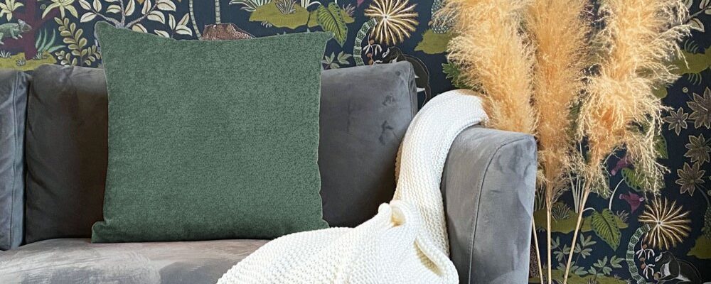 A cushion in snug, moss green colour, providing a pop of solid colour against the dark background. A sofa in a soft, dark grey or charcoal velvet/suede-like fabric which the cushion is resting on.