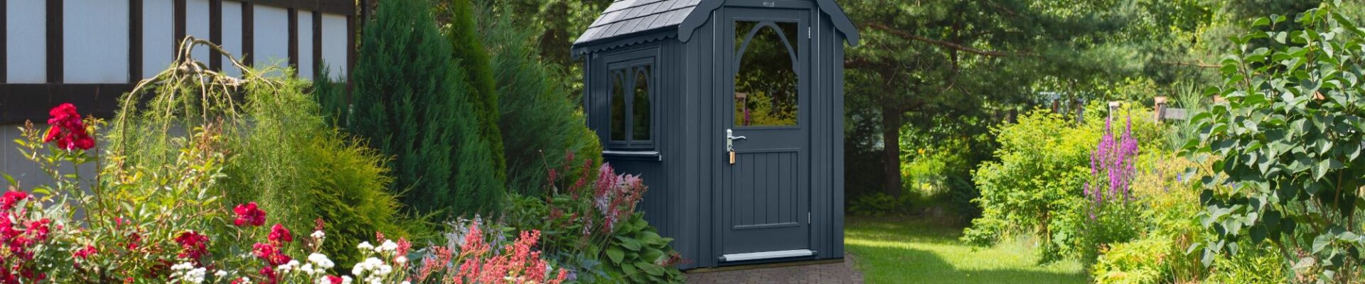An ornate garden shed in a dark grey colour. It sits in a beautifully manicured garden.
