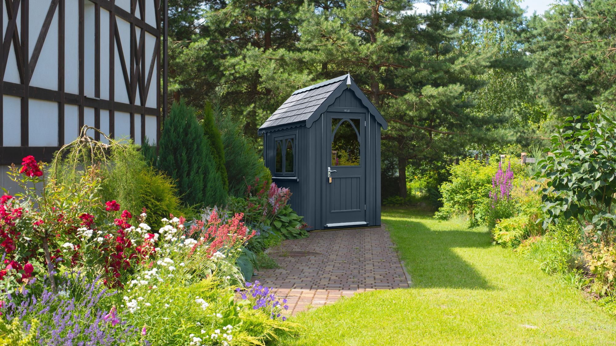 An ornate garden shed in a dark grey colour. It sits in a beautifully manicured garden.