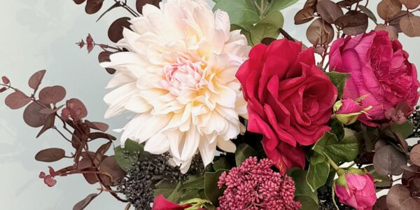 A close-up of an artificial flower bouquet featuring a large, creamy white dahlia, a deep red rose, and a magenta peony, surrounded by burgundy eucalyptus leaves and small dark berries.