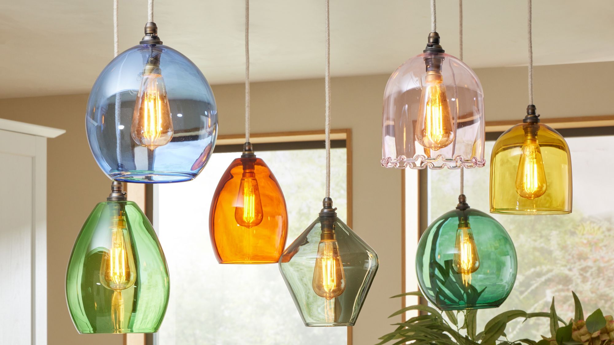 The image shows a cluster of seven colourful glass pendant lights hanging from the ceiling, likely over a kitchen island or dining area. Each pendant has a distinct shape and colour, creating a vibrant, eclectic look.