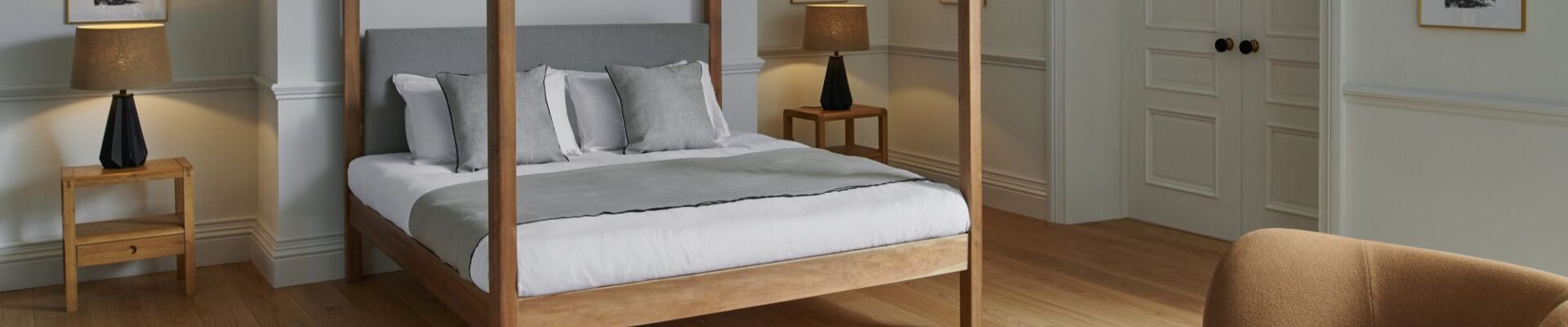 A bright, contemporary bedroom with light hardwood floors and pale grey walls. The centrepiece is a wooden four-poster bed with a grey upholstered headboard, white linens, and a grey throw blanket, accented by several pillows in white and light grey.