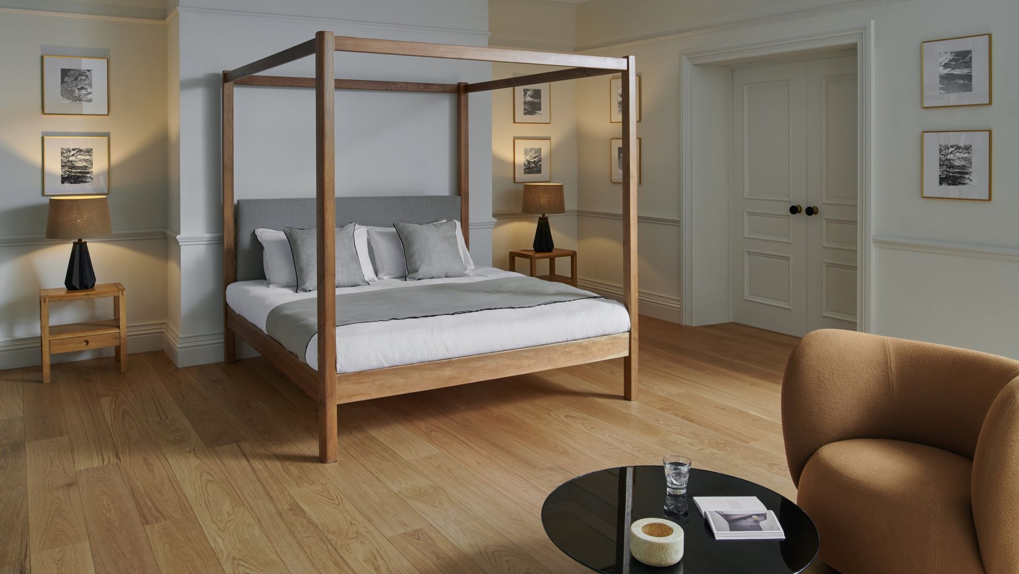 A bright, contemporary bedroom with light hardwood floors and pale grey walls. The centrepiece is a wooden four-poster bed with a grey upholstered headboard, white linens, and a grey throw blanket, accented by several pillows in white and light grey.