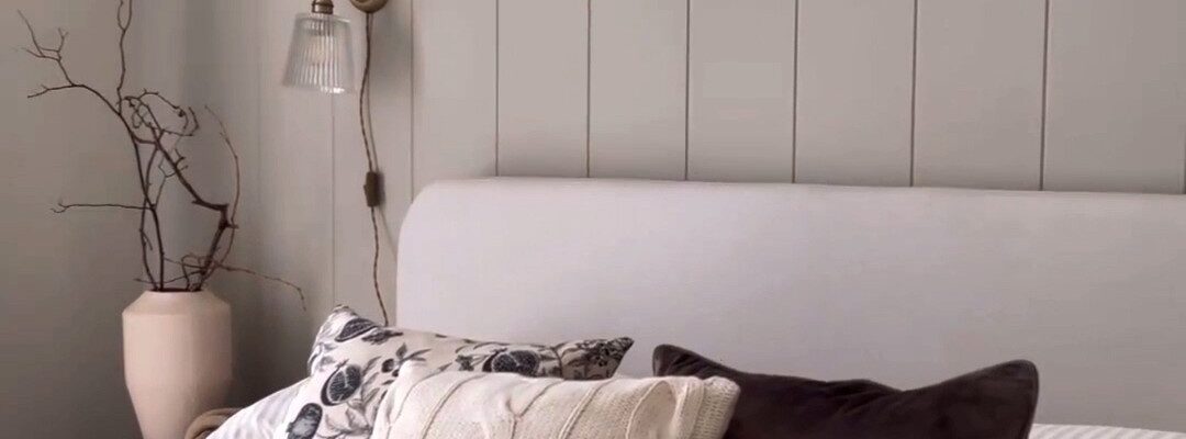 The wall features vertical V-groove panelling painted a light, neutral grey/taupe colour. The panelling extends upwards, perhaps to meet a picture rail or just below the ceiling, creating a backdrop for the bed.