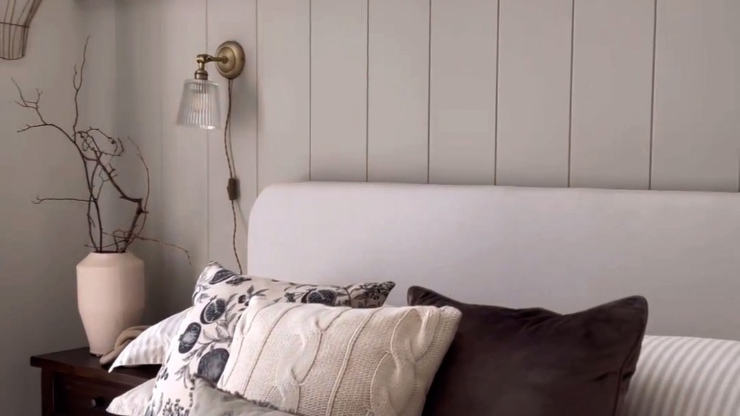 The wall features vertical V-groove panelling painted a light, neutral grey/taupe colour. The panelling extends upwards, perhaps to meet a picture rail or just below the ceiling, creating a backdrop for the bed.