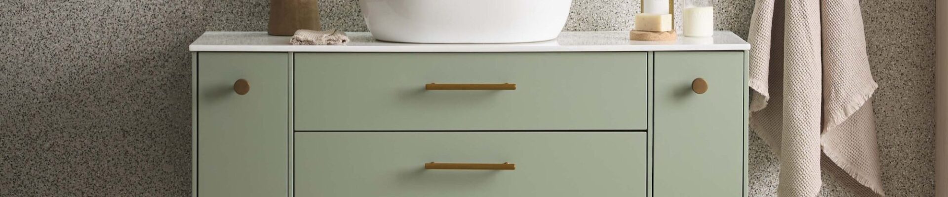 A wall-mounted vanity in a soft, muted sage green or light olive colour. It has two central drawers with long, horizontal brass or gold-tone handles.