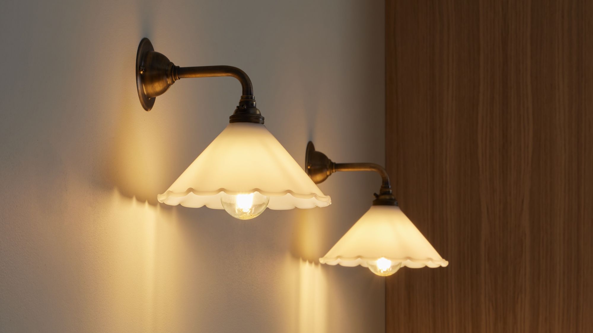 Two contemporary brass wall lights with opal frilled glass shades mounted on a white wall.