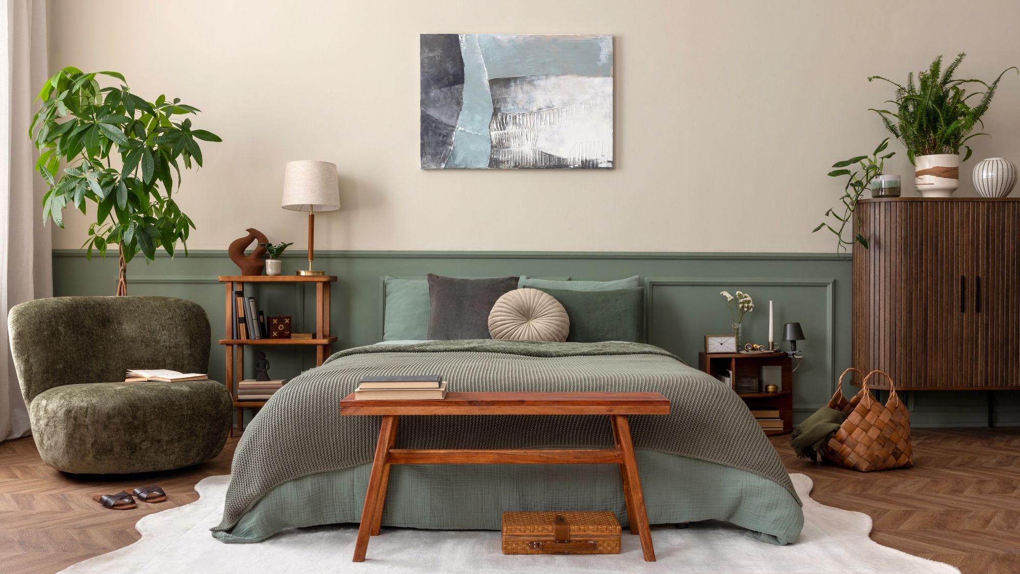 A cosy, biophilic-style bedroom featuring a bed with sage green linens and a textured moss-green throw. The wall behind the bed is split into a warm oatmeal top and muted green wood panelling on the bottom.
