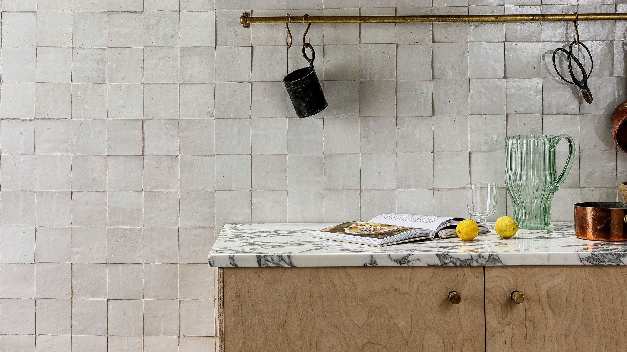 A kitchen interior featuring a handmade white Zellige tile backsplash with a textured, uneven surface. A marble countertop sits atop light wood cabinetry, holding an open cookbook, two lemons, and a green fluted glass pitcher. Above, a brass rail hangs against the tile with kitchen tools and a small black cup.