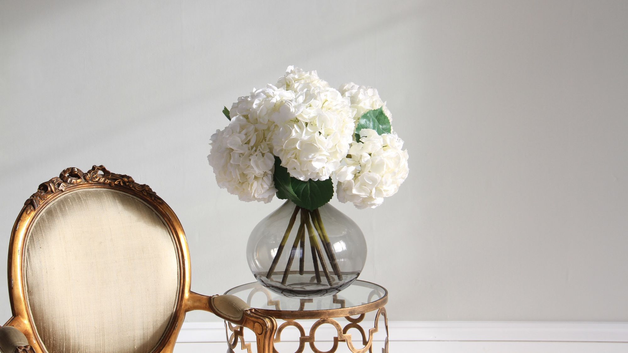 : An elegant indoor setup featuring a bouquet of artificial white hydrangeas in a round, smoky-grey glass vase. The vase sits on a small, gold-finished side table with a circular glass top and ornate geometric detailing.