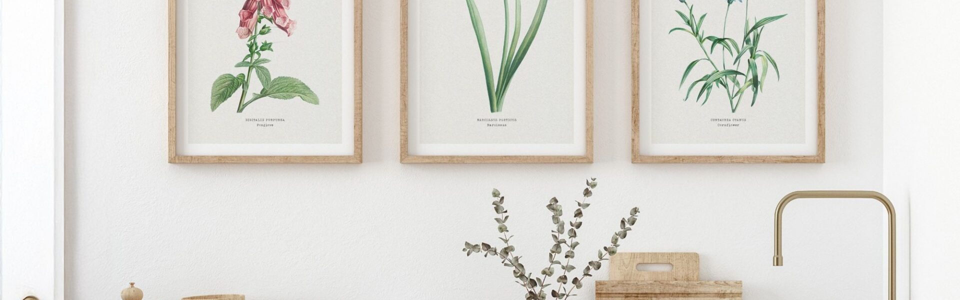 A trio of framed botanical art with Foxglove, Daffodils, and Cornflower prints hangs above a well-appointed light wood kitchen counter featuring assorted bowls, kitchen tools and a vase of eucalyptus.