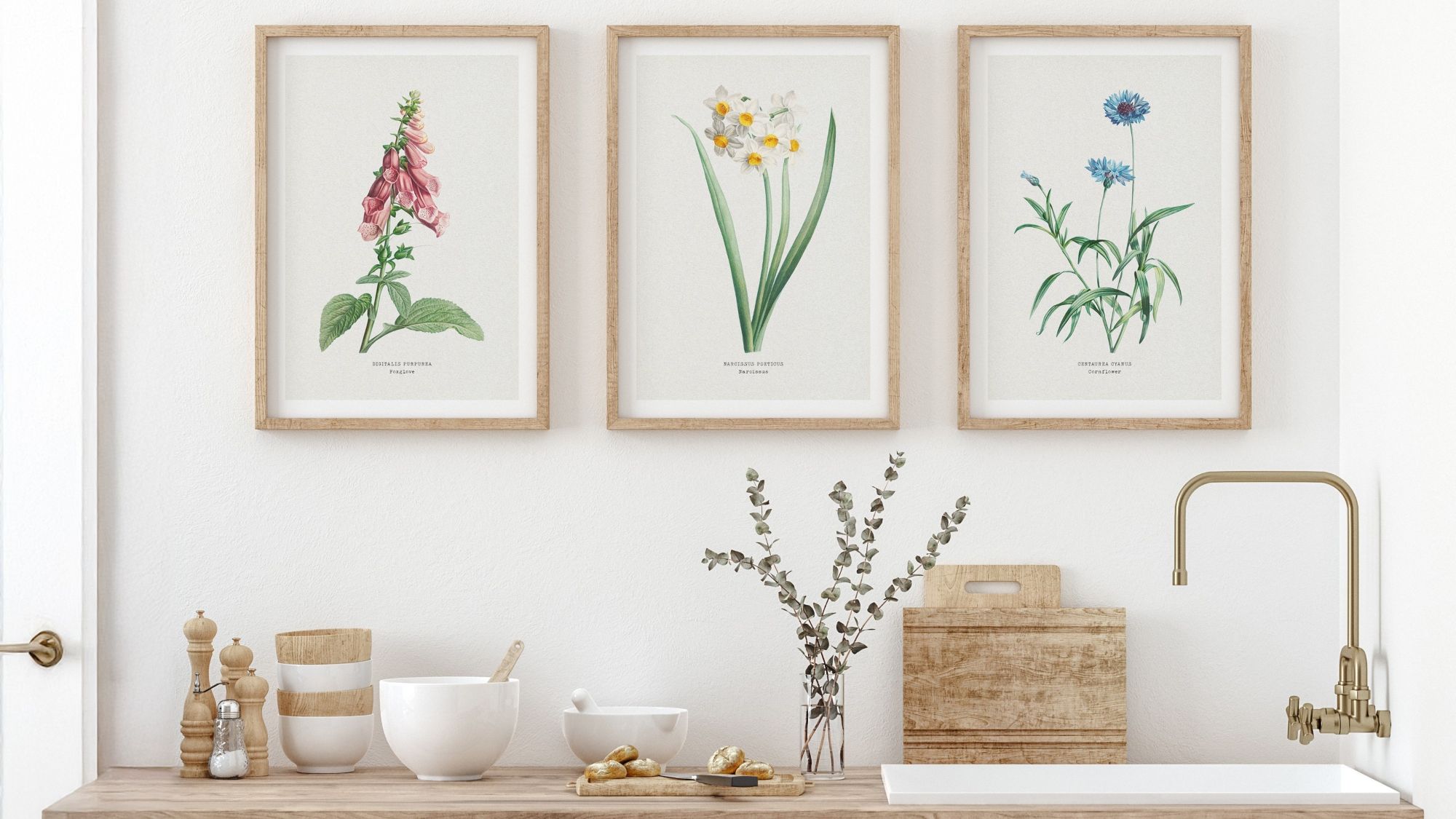 A trio of framed botanical art with Foxglove, Daffodils, and Cornflower prints hangs above a well-appointed light wood kitchen counter featuring assorted bowls, kitchen tools and a vase of eucalyptus.