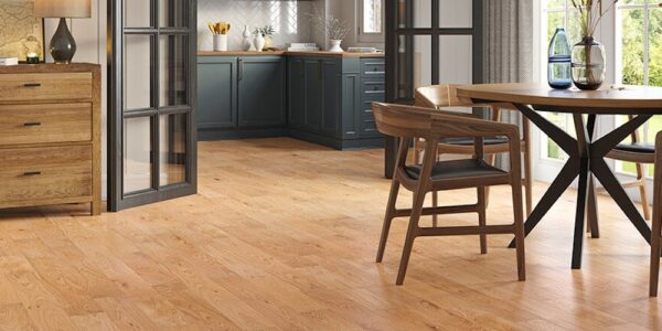 A modern open-plan kitchen and dining area featuring warm Lusso Carrara Orchard engineered oak flooring, dark teal cabinetry, and mid-century modern wooden furniture.