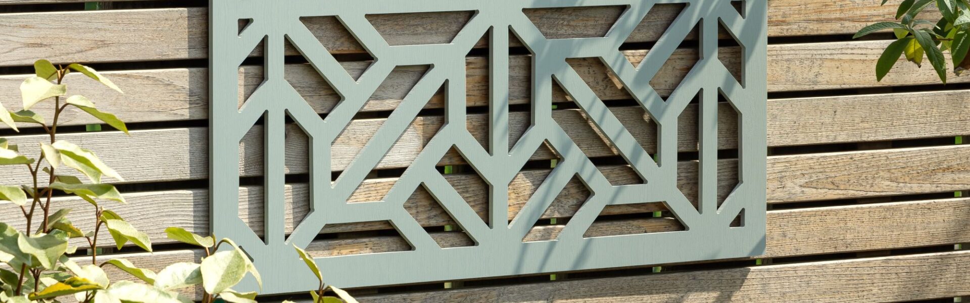 A decorative, square, sage-green wooden privacy screen with an intricate geometric, star-shaped cut-out pattern is mounted on a light-coloured, slatted wooden fence. The screen is centred in the upper portion of the frame, showcasing the detailed symmetry of the cutouts against the fence's wood grain.