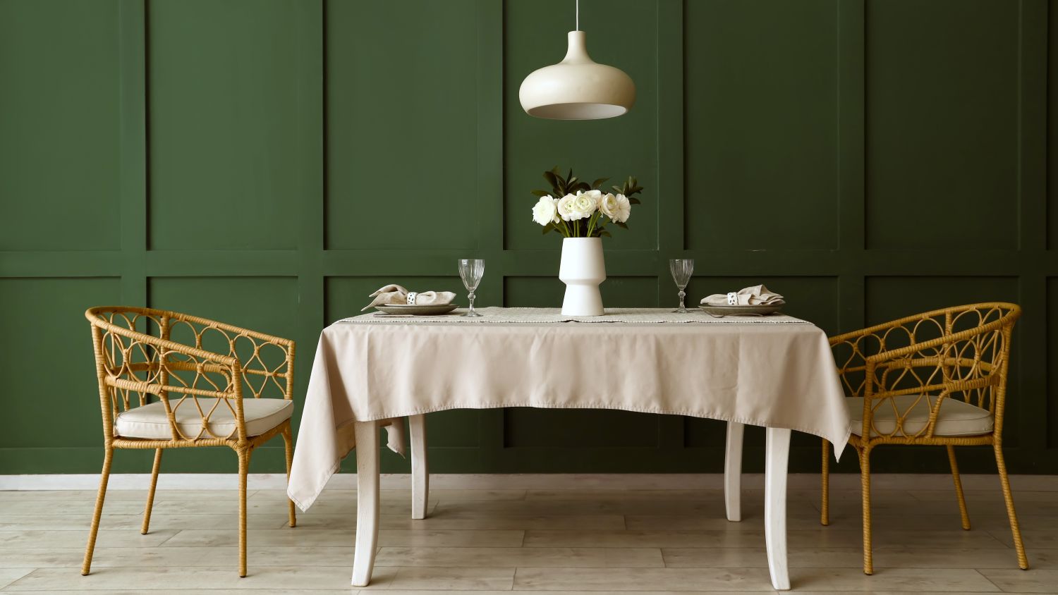 An elegant, eye-level wide shot of a dining area featuring a deep pepper tree green wainscoted wall. Centred in the room is a rectangular white wooden dining table draped with a light beige tablecloth, set with two minimalist place settings, crystal wine glasses, and a white vase holding white ranunculus flowers.