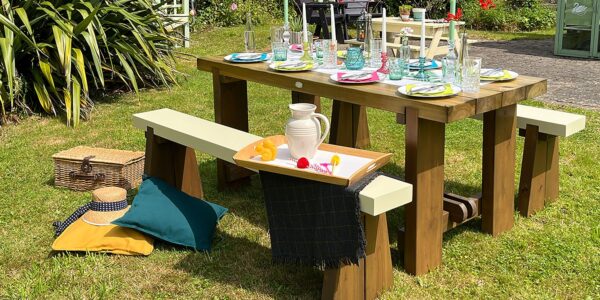 An outdoor wooden dining set sits on a lush green lawn on a sunny day. The set features a heavy, stained wooden table and two matching benches with contrasting cream-colored tops.