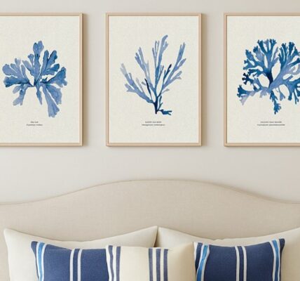 A wide landscape shot of a coastal-themed interior featuring a set of three botanical seaweed art prints hanging on an off-white wall. Below the artwork is a vibrant olive-green wainscoted wall, and in the foreground, a vintage-style brass bathtub faucet and shower head are partially visible, suggesting a sophisticated, spa-like bathroom setting. Soft, natural sunlight casts gentle shadows across the scene from the right.