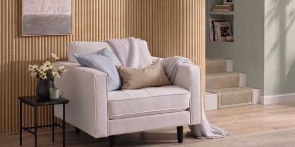 A cosy, modern white armchair sits in front of a light oak reeded wood-panelled feature wall. The armchair is styled with a light blue cushion, a tan striped lumbar pillow, and a soft lavender throw blanket.