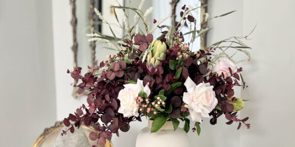 A lush autumn floral arrangement in a matte white ceramic vase, featuring deep plum-coloured eucalyptus foliage, pale pink roses, small pink berries, a green protea bud, and wispy ornamental grasses, set against a bright interior with a gold-framed mirror in the background.