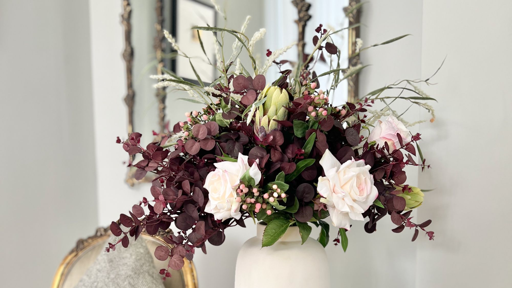 A lush autumn floral arrangement in a matte white ceramic vase, featuring deep plum-coloured eucalyptus foliage, pale pink roses, small pink berries, a green protea bud, and wispy ornamental grasses, set against a bright interior with a gold-framed mirror in the background.