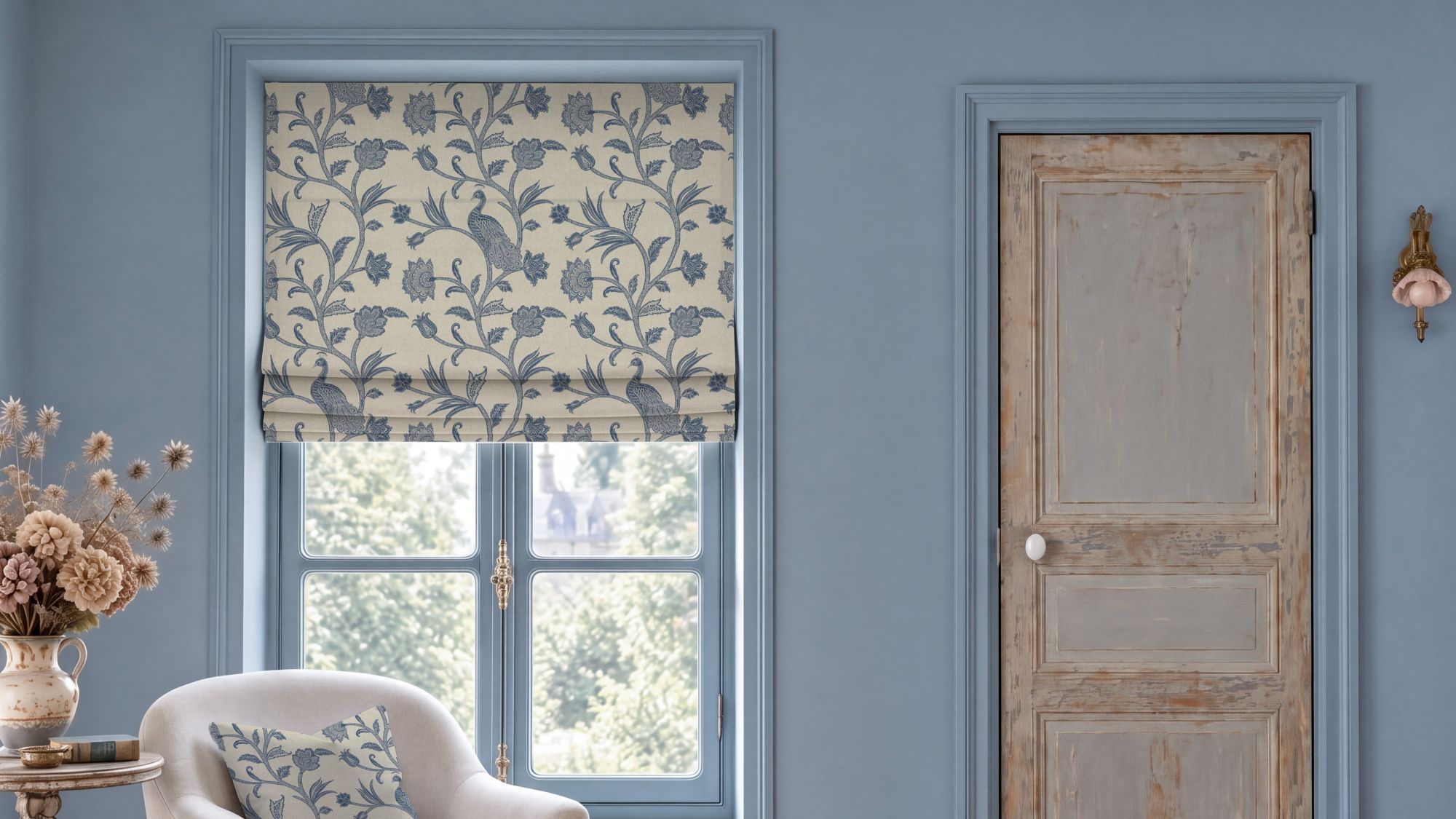 Escape to the Chateau L'Oiseau du Chateau Roman blind in blue featuring a vintage botanical bird and floral pattern, hung over a window in a blue room with a distressed wooden door