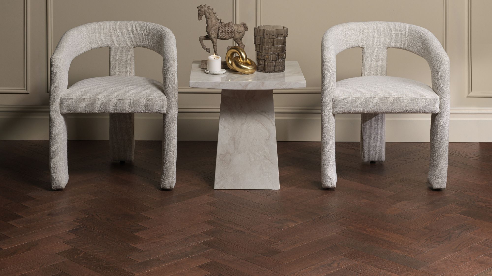 Two light grey upholstered accent chairs set against wainscoted walls, featuring a small marble side table between them on dark brown oak herringbone parquet flooring.