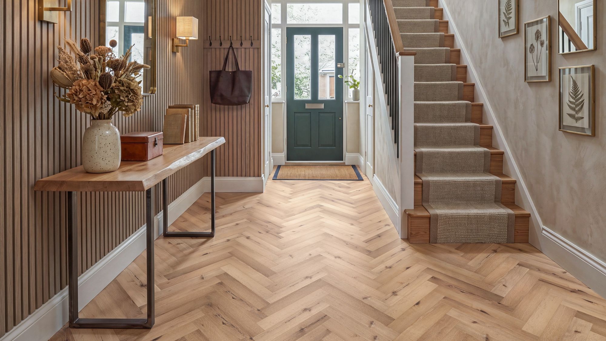 Bright and stylish hallway featuring burnished oak herringbone LVT flooring, a live-edge console table with dried florals, slatted wood accent walls, and a staircase with a textured runner leading to a dark green front door