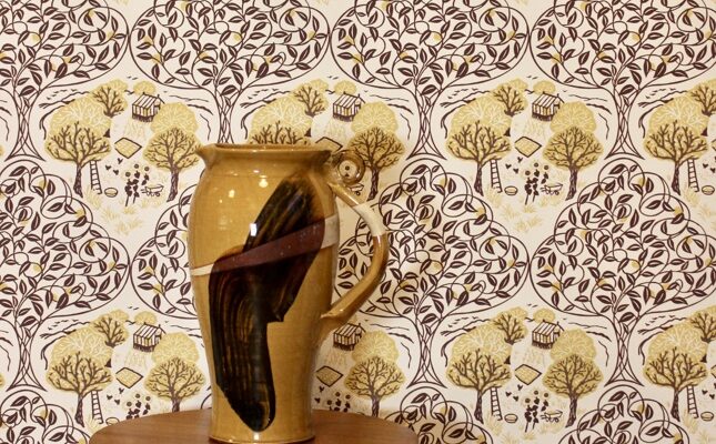 A wallpaper behind a vase which depicts a charming countryside scene and is available in seven colourways.